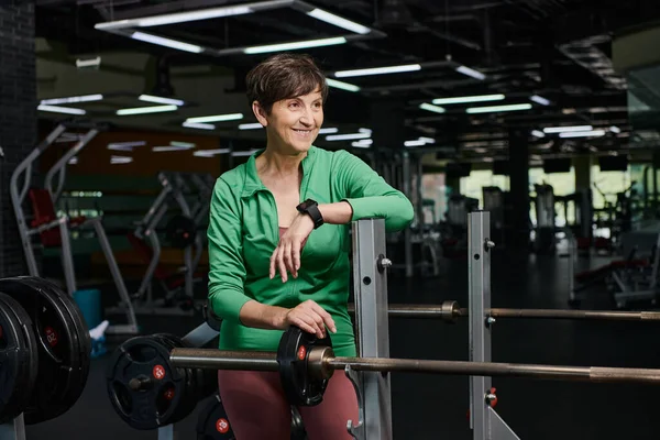 Felice donna anziana sorridente e in piedi vicino al bilanciere, dischi, allenamento in palestra, motivazione — Foto stock