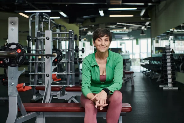Glückliche ältere Frau lächelt und sitzt neben Langhantel, Hantelscheiben, Training im Fitnessstudio, Motivation — Stockfoto