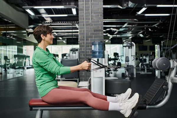 Atlético e motivado, mulher idosa trabalhando no ginásio, fitness, máquina de exercícios, vista lateral — Fotografia de Stock