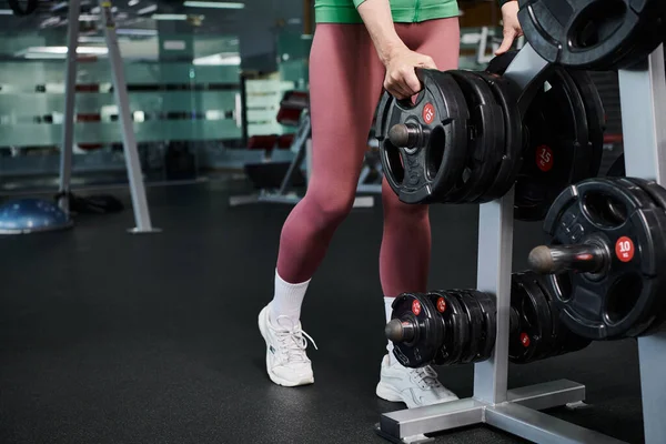 Esportista cultivado em leggings e tênis branco de pé perto barbell no ginásio, placas de peso — Fotografia de Stock