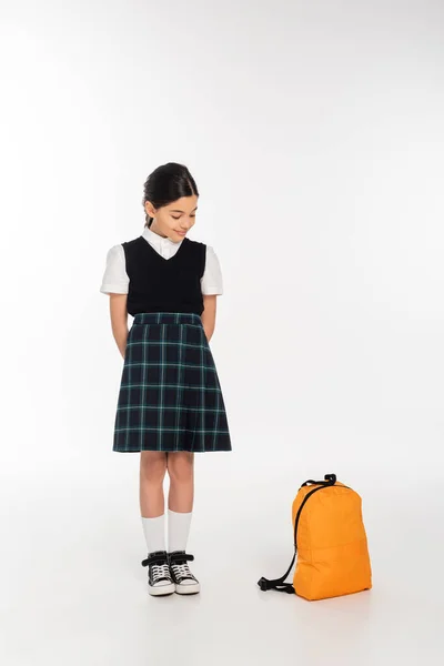 Menina feliz no uniforme da escola de pé e olhando para a mochila no fundo branco, de volta à escola — Fotografia de Stock