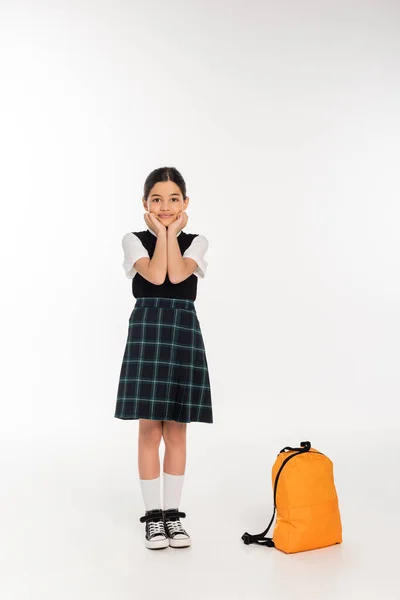 Menina feliz no uniforme da escola de pé perto da mochila no fundo branco, de volta à escola, comprimento total — Fotografia de Stock
