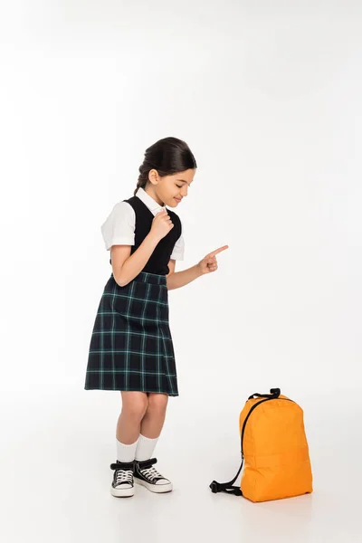 Fille heureuse en uniforme scolaire debout et regardant sac à dos sur fond blanc, pointant du doigt — Photo de stock
