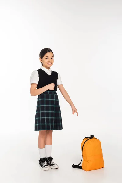 Menina alegre em uniforme escolar de pé e apontando para a mochila no fundo branco, comprimento total — Fotografia de Stock