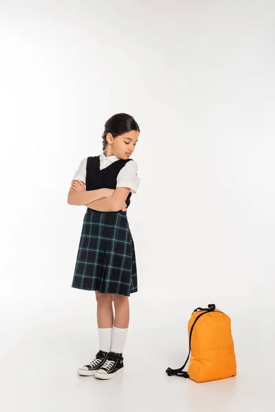 Displeased schoolgirl standing with folded arms and looking at backpack, full length, school concept — Stock Photo