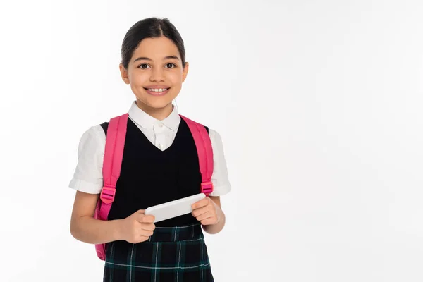 Âge numérique, heureuse écolière avec sac à dos tenant smartphone isolé sur blanc, étudiante en uniforme — Photo de stock