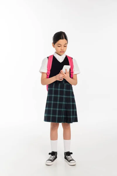 Цифровой возраст, школьница с рюкзаком держа смартфон на белом, студент в форме, полная длина — стоковое фото