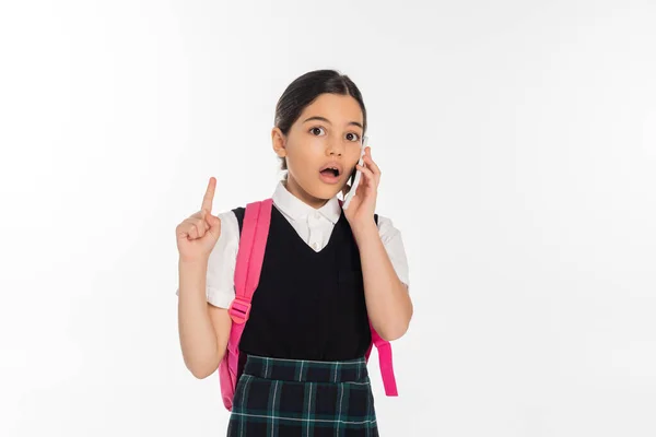 Digitales Zeitalter, staunendes Schulmädchen mit Rucksack spricht auf Smartphone isoliert auf weiß und zeigt nach oben — Stockfoto