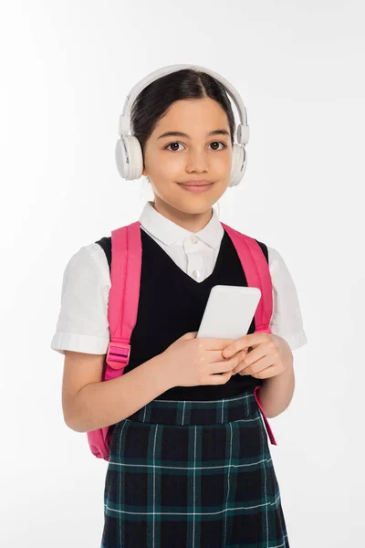 Цифровий вік, радісна школярка в бездротових навушниках тримає смартфон ізольовано на білому, студент — стокове фото