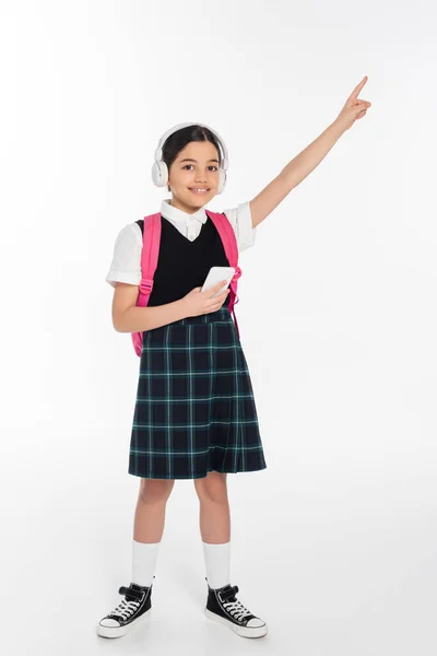 Digital age, cheerful schoolgirl in wireless headphones holding smartphone on white, pointing away — Stock Photo