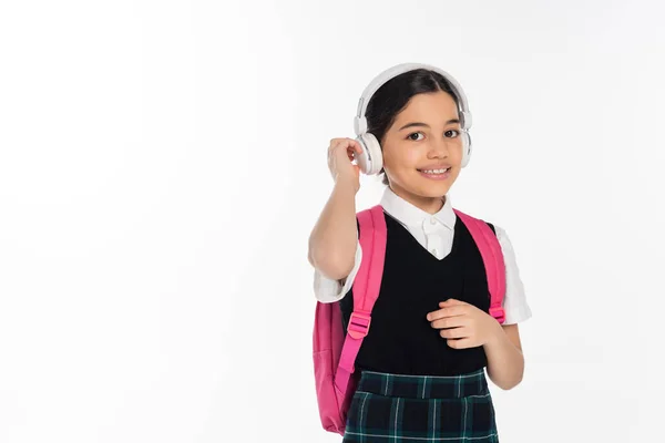 Music, cheerful schoolgirl in wireless headphones looking at camera isolated on white, student — Stock Photo