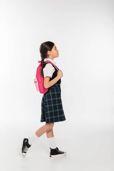 Longitud completa, colegiala en uniforme de pie con mochila y mirando hacia otro lado, fondo blanco - foto de stock