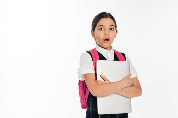 Impactado colegiala sosteniendo el ordenador portátil y mirando a la cámara, chica en uniforme escolar, aislado en blanco - foto de stock