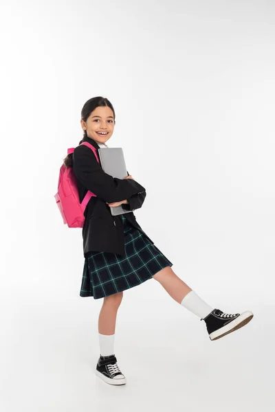 Alegre colegiala sosteniendo portátil y mirando a la cámara, chica en uniforme escolar sobre fondo blanco - foto de stock