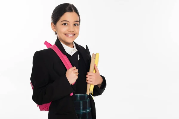 Веселая школьница, стоящая с блокнотами и рюкзаком, изолированная на белом, возвращаясь к школьной концепции — стоковое фото