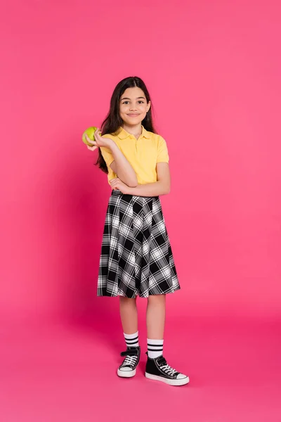 Happy brunette schoolgirl holding fresh apple on pink background, bright and vibrant, plaid skirt — Stock Photo