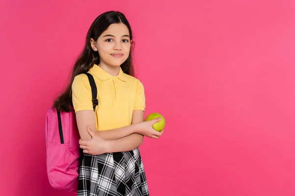 Щаслива школярка з рюкзаком тримає зелене свіже яблуко ізольовано на рожевому, яскравому фоні — стокове фото