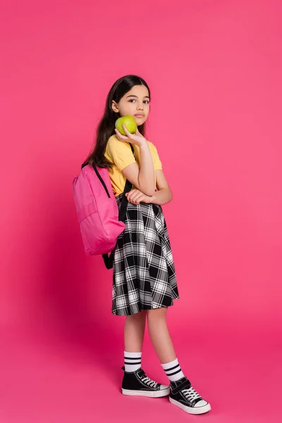Volle Länge, brünettes Schulmädchen steht mit Rucksack und hält grünen Apfel, rosa Hintergrund — Stockfoto
