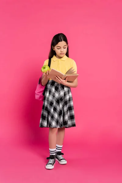 Full length, brunette schoolgirl holding green apple and reading book on pink background, backpack — Stock Photo