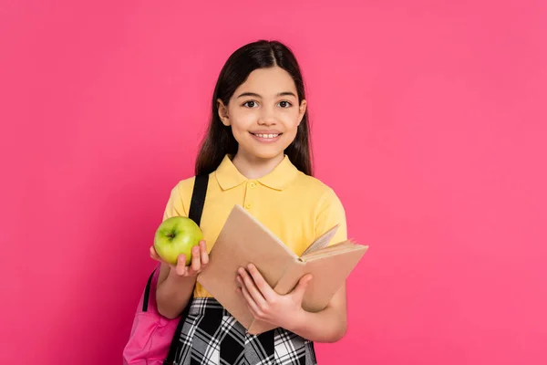 Joy, brunette schoolgirl holding green apple and reading book isolated on pink, girl with backpack — Stock Photo