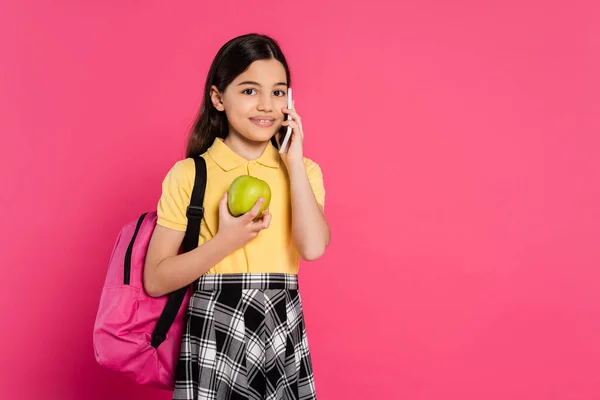 Colegiala feliz de pie con la mochila, la celebración de manzana y hablar en el teléfono inteligente, llamada telefónica - foto de stock