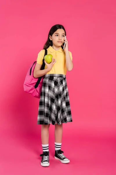 Брюнетка школярка говорить на смартфоні на рожевому фоні, телефонний дзвінок, яблуко і рюкзак — стокове фото