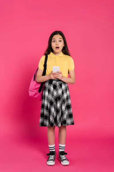 Chica asombrada, colegiala sosteniendo teléfono inteligente y mirando a la cámara en el fondo rosa, vibrante - foto de stock