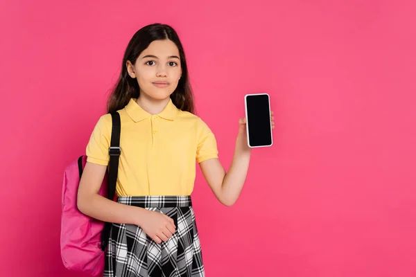 Colegiala morena sosteniendo smartphone con pantalla en blanco y mirando a la cámara sobre fondo rosa - foto de stock