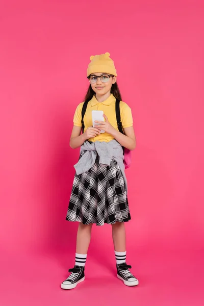 Chica feliz en gorro sombrero y gafas usando teléfono inteligente sobre fondo rosa, longitud completa, mochila - foto de stock