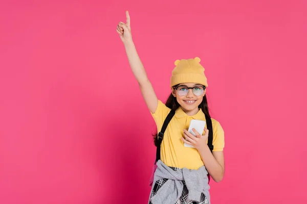Menina feliz em chapéu gorro e óculos usando smartphone no fundo rosa, olhar elegante, apontando para cima — Fotografia de Stock