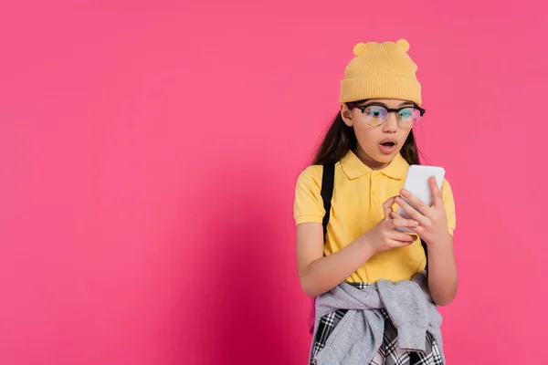 Écolière choquée en bonnet chapeau et lunettes à l'aide d'un smartphone sur fond rose, look élégant — Photo de stock