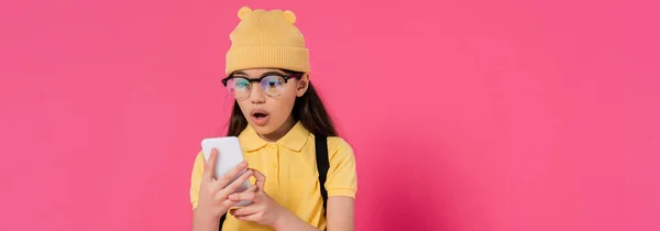 Shocked schoolgirl in beanie hat and glasses using smartphone on pink background, stylish, banner — Stock Photo