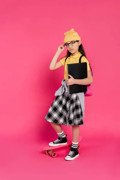 Schoolgirl in beanie hat and glasses holding notebooks, standing near penny board on pink background — Stock Photo