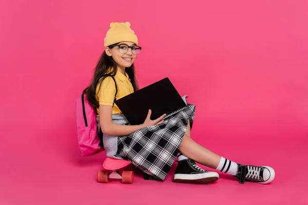Happy schoolgirl in beanie hat and glasses sitting on penny board, pink background, notebooks — Stock Photo