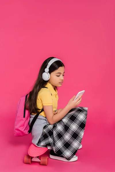 Colegiala en auriculares inalámbricos sentado en el tablero de penique, fondo rosa, utilizando el teléfono inteligente - foto de stock