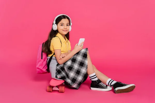 Colegiala feliz en los auriculares sentados en el tablero del centavo, fondo rosa, usando el teléfono inteligente, digital - foto de stock