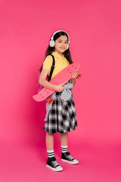 Alegre chica en auriculares inalámbricos de pie con penny board, fondo rosa, después de clases - foto de stock