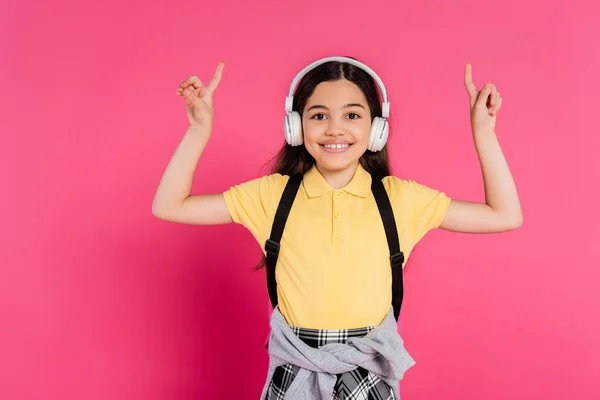 Chica positiva en auriculares inalámbricos que muestran signo v sobre fondo rosa, emoción, después de clases - foto de stock