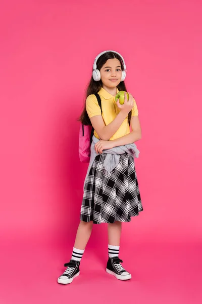Estudante sorrindo em fones de ouvido sem fio segurando maçã verde no fundo rosa, criança com mochila — Fotografia de Stock