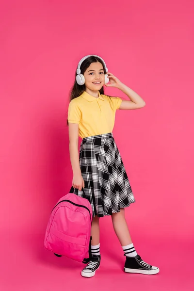 Studentessa eccitata in cuffie wireless tenendo lo zaino su sfondo rosa, studente bruna — Foto stock