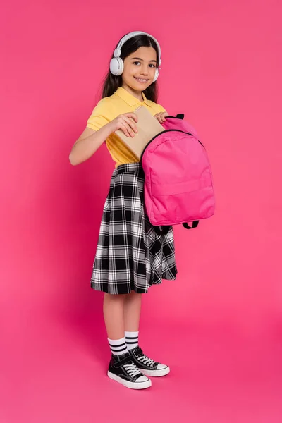 Happy schoolgirl in wireless headphones putting book inside of backpack, pink background, student — Stock Photo