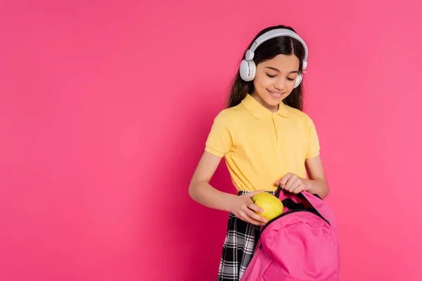 Happy schoolgirl in wireless headphones putting apple inside of backpack, pink background, student — Stock Photo
