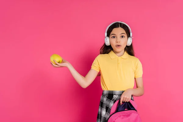 Colegiala conmocionada en auriculares inalámbricos con manzana y mochila, fondo rosa, estudiante - foto de stock