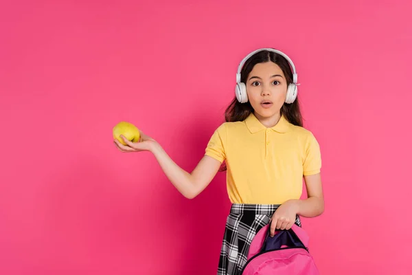 Colegiala conmocionada en auriculares inalámbricos con manzana y mochila, fondo rosa, estudiante - foto de stock