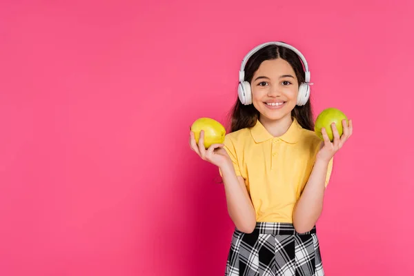 Colegiala sorprendida en auriculares inalámbricos sosteniendo manzana y mochila, fondo rosa, estudiante - foto de stock