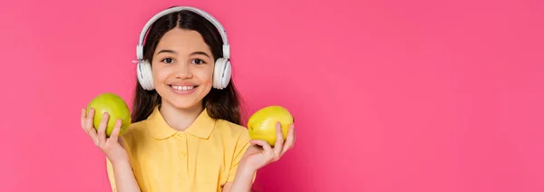 Positive schoolgirl in wireless headphones holding green apples on pink background, brunette student — Stock Photo