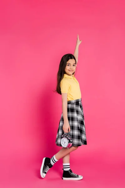 Glückliches Schulmädchen hält Wecker auf rosa Hintergrund, blickt in die Kamera, zeigt nach oben, volle Länge — Stockfoto