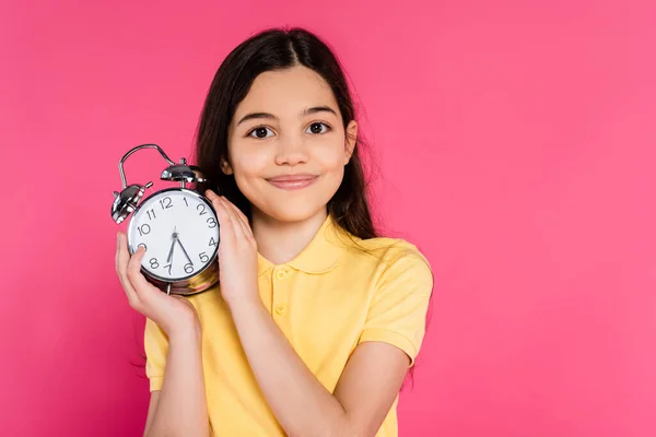 Retrato, colegiala feliz sosteniendo reloj despertador aislado sobre fondo rosa, mirando a la cámara - foto de stock