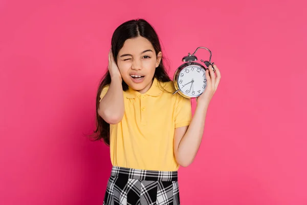 Colegiala mirando a la cámara, guiñando el ojo, sosteniendo reloj despertador vintage aislado en rosa, de vuelta a la escuela - foto de stock