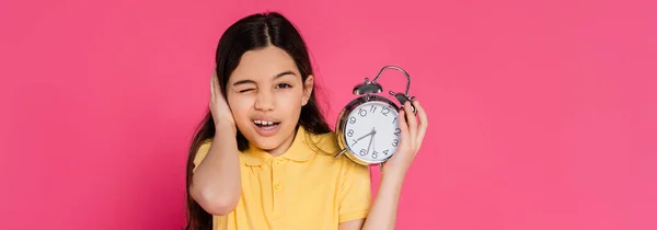 Schoolgirl looking at camera, winking, holding alarm clock isolated on pink, back to school, banner — Stock Photo